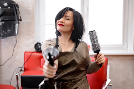 a black-haired woman hairdresser sits in a hairdresser's chair and laughs as she holds a brushing and hairdryerの写真素材