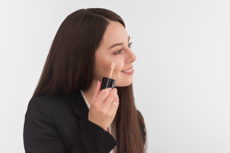 girl, make-up artist, with long dark hair in a business suit, applies a concealer on the face on a white backgroundの写真素材