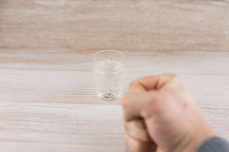 the struggle with addiction - alcoholism: a shot of vodka on the table in front of a man, clenched fist, in search of sufficient strength of will to resist temptationの写真素材