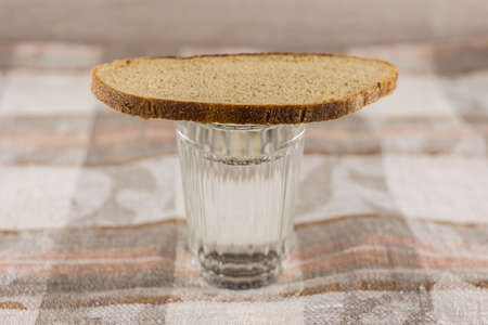 Russian symbol of commemoration of the dead: a loaf of dark rye bread on a full glass of Russian vodka. Wooden table white colors, front viewの写真素材