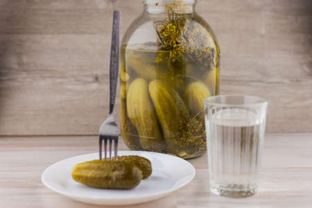 national trait harmful to health: a large jar of pickles and faceted glass of Russian vodka on a white table, a couple of cucumbers laying on the plate, a fork sticking in it, front viewの写真素材