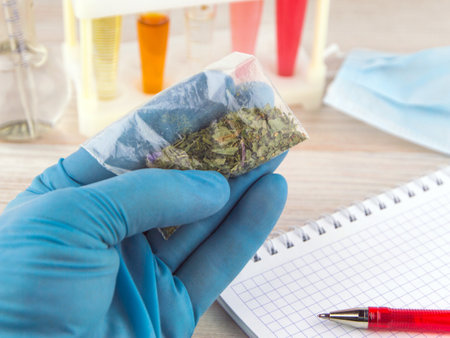 chemical analysis of "spice": a bag with a synthetic Smoking mixture is considered by a person in the hand wearing a blue glove against the background of tubes and a notebook and a bulb with a pipetteの写真素材