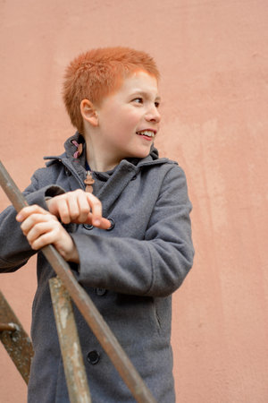 red-haired boy in a gray coat climbed the rusty metal ladder to the wall and happily look backの写真素材