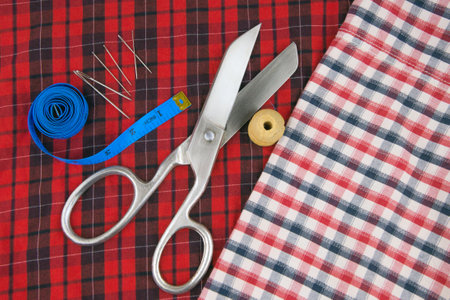 sewing tools: large scissors and thread with needles lying on a checkered red cloth in the blue measuring tape, white plaid fabric leftの写真素材