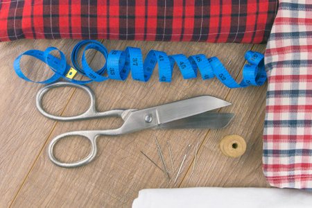 sewing tools: large scissors, measuring tape, spool of thread and needles lie side by side on a wooden table, different fabric lying aroundの写真素材