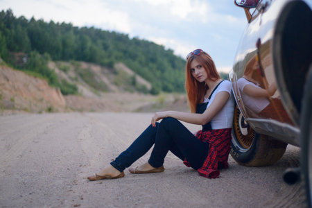girl in sunglasses sitting on the pavement near the red car and looks at us quietly, hills with green forest in the backgroundの写真素材