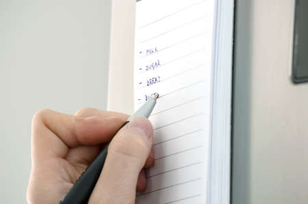 Female hand writing grocery list on a sticky reminder note on a refrigerator の写真素材
