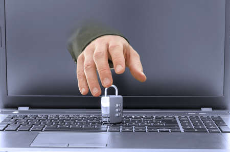 Closeup of hacker hand coming out of laptop screen trying to steal or break a padlock on keyboard.