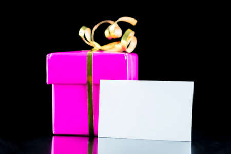Pink wrapped gift box wit golden bow and blank greeting card. Isolated over black background.