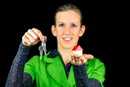 Young female real estate agent holding wooden toy house in one and keys in other hand. Isolated over black background.の写真素材