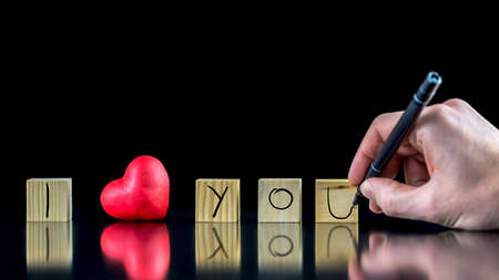 Valentines concept with a man writing with a pen on wooden blocks - I Love You - with a symbolic red heart depicting his Love and commitmentの写真素材