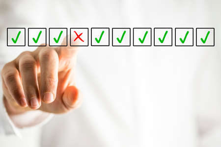 Man selecting a red cross in a box from a line of green check marks or ticks in boxes on a virtual screen conceptual of individuality, quality, failure and negativityの写真素材