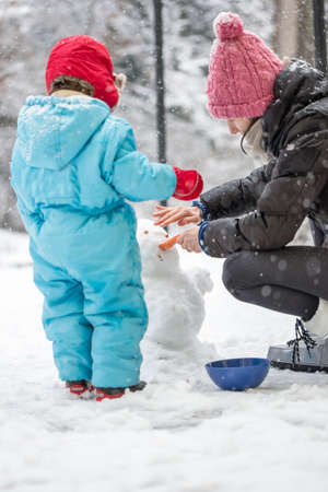 View from the rear of a young mother and toddler working in the fresh white snow in the garden building a winter snowmanの写真素材