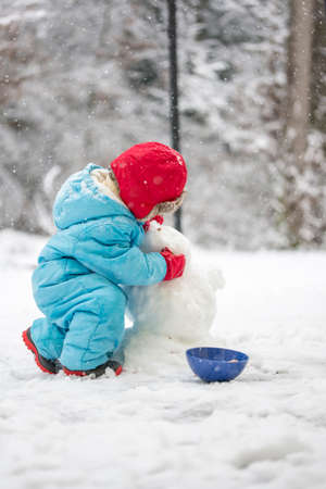 Young child building a snowman wrapped up warmly against the cold in a blue jumpsuit bending down in the cold white winter snow shaping the head, view from behindの写真素材
