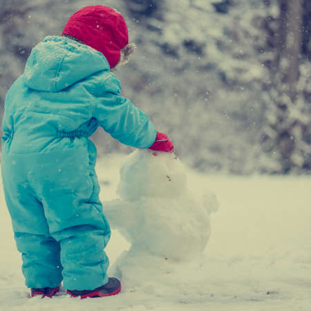 Cute young toddler in a warm winter outfit standing in a cold wintry garden building a snow man from...