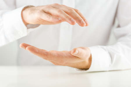 Man cupping his hands in a protective gesture above and below an empty space for your product placement or conceptual object, close up view of the hands against a white shirt.の写真素材