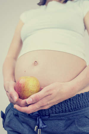 Low angle toned retro effect image of a pregnant woman holding an apple near her swollen bare belly conceptual of healthy eating and diet during pregnancy.の写真素材