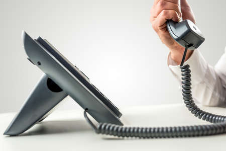 Side view of a black business landline telephone with the receiver held by a male hand with white...