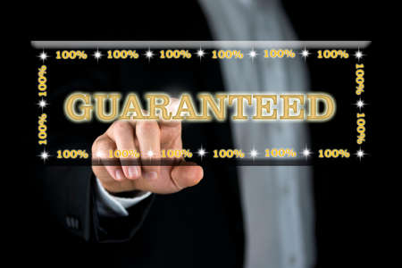 Businessman activating a navigation bar on a virtual screen with his finger containing the words - Guaranteed 100 percent - in gold lettering in a conceptual image, close up of the button and his handの写真素材