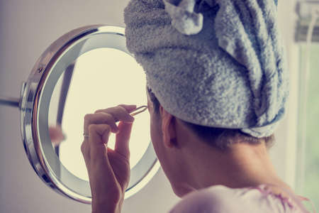 Retro instagram style image of a woman with her hair wound in a towel carefully plucking her...