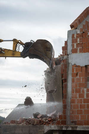 Bucket of a backhoe or mechanical digger against the skyline demolishing the wall of a brick building with flying masonry and debris.の写真素材