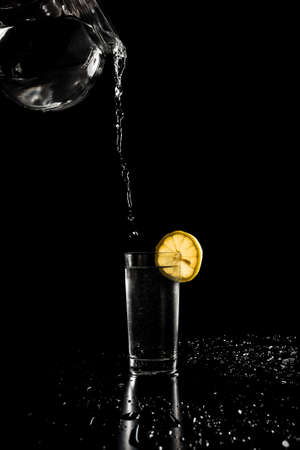 Pouring water in to a tall glass garnished with a slice of fresh lemon from a glass jug held at a height with spillage around the glass on a dark background with copyspace.の写真素材