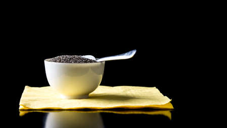 Bowl of healthy chia seeds rich in omega-3 fatty acids standing with a spoon on a black reflective table against a black background with copyspace.の写真素材