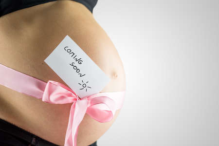 Young woman in the last stages of pregnancy with a pink bow tied around her swollen bare abdomen or baby bump with a handwritten note - coming soon - side view on grey with copyspace.の写真素材