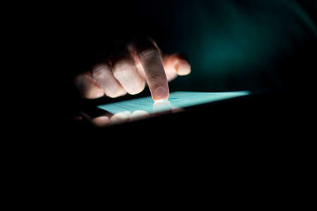 Man using a tablet in the dark navigating the glowing blue touchscreen with his finger , with copyspace.の写真素材