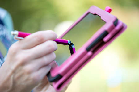 Close up view of the hand of a woman standing outdoors writing on the touch screen of a pink tablet...