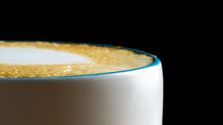 Low angle view across the surface of the bubbles of a freshly brewed cup of frothy cappuccino coffee garnished with cream over a black background.の写真素材