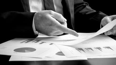 Businessman analysing a set of bar, pie and triangular graphs as he sits at his desk pointing to one document he is holding, close up greyscale in a business analysis, projections and strategy conceptの写真素材