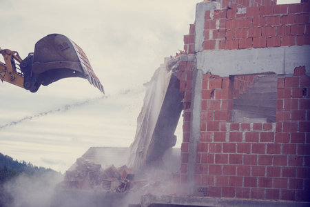 Retro image of bucket of a backhoe or mechanical digger demolishing the wall of a brick building with flying masonry and debris.の写真素材