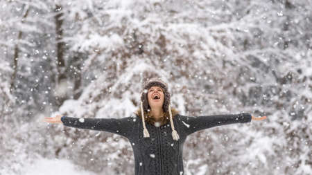 Happy vivacious woman celebrating the winter snow standing with her arms outspread in a snowy forest laughing as she watches the falling snowflakes from above.の写真素材