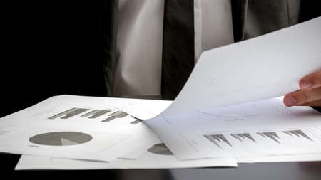 Close up of the hands of a businessman seated at his desk analysing a set of statistic report and...