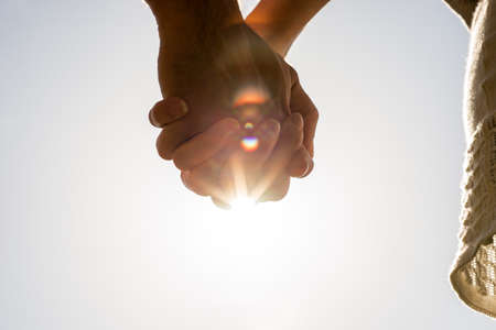 Clasped hands of a young romantic man and woman against a bright sun flare with copyspace, conceptual image of love and friendship.の写真素材