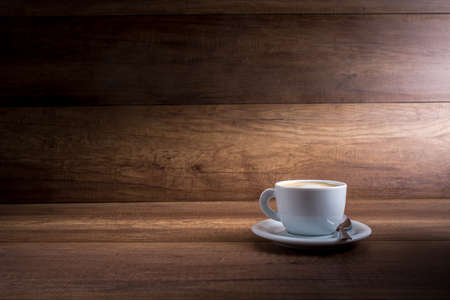 Cup of freshly brewed aromatic cappuccino standing on a wooden table in a shaft of light against a rustic wood background with plenty of copyspace.の写真素材
