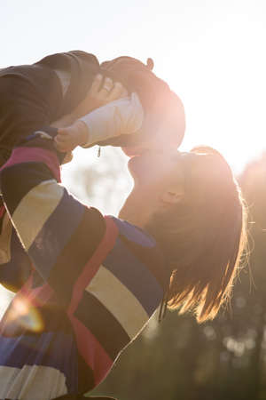 Joyful young mother with her young baby kissing the child as she holds it up in the air in front of the warm glow of a morning sunburst.の写真素材