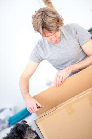 Man in casual clothes kneeling on the floor unpacking a flat cardboard carton, tilted angle close up view.の写真素材
