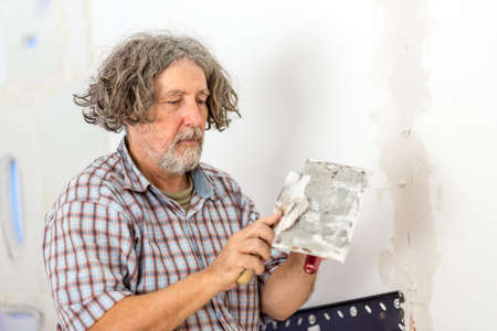 Middle-aged male builder or homeowner repairing a wall applying filler plaster from a small board...