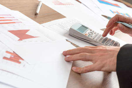 Businessman analysing statistical graphs using a manual calculator at his desk in the office, close...