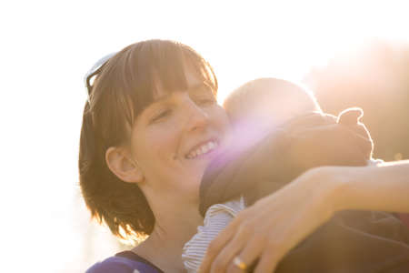 Happy young  protective mother lovingly cuddling her baby boy to her shoulder with maternal smile backlit by a bright sunburst.の写真素材