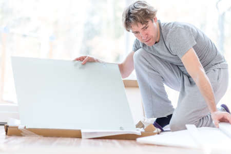Man unpacking flat pack components or panels of an item of white furniture as he kneels on the floor in the living room of a house.の写真素材