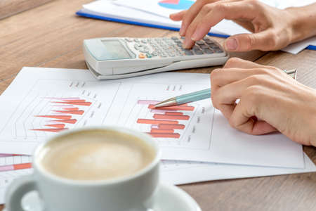 Female accountant working on a presentation or annual report analysing a set of graphs and charts with a manual calculator with a cup of coffee alongside.の写真素材