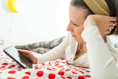 Young woman wearing a golden alice band in her hair looking on digital tablet or e-book device lying on her bed on a red and white polka dot bedspread.の写真素材