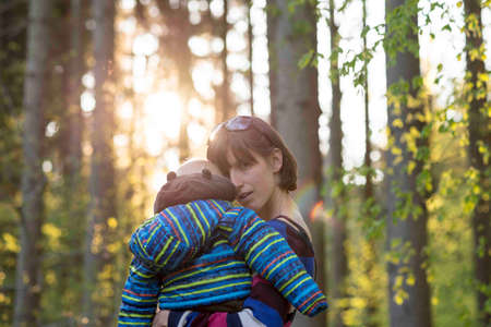 Loving mother carrying her young child through the woods at sunrise with the rising sun behind her head as she nuzzles her toddler in an intimate embrace.の写真素材
