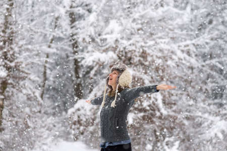 Happy young woman embracing beautiful life with her arms wide open as she is standing outside in a snowy winter woodland with snowflakes falling.の写真素材
