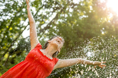 Happy young  woman standing under a spray of water on a hot summer day with her arms outstretched rejoicing, celebrating and enjoying life, leafy green tree background.の写真素材