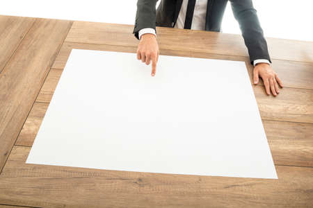 Businessman leaning over a wooden desk pointing to a blank white placard or large sheet of paper in a presentation and planning concept, high angle view of his hand and desk.の写真素材