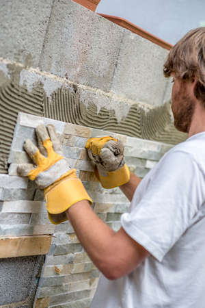 Builder tiling a wall with ornamental tiles pushing the tile into the cement on the wall with his gloved fist in a DIY, renovation or construction conceptの写真素材